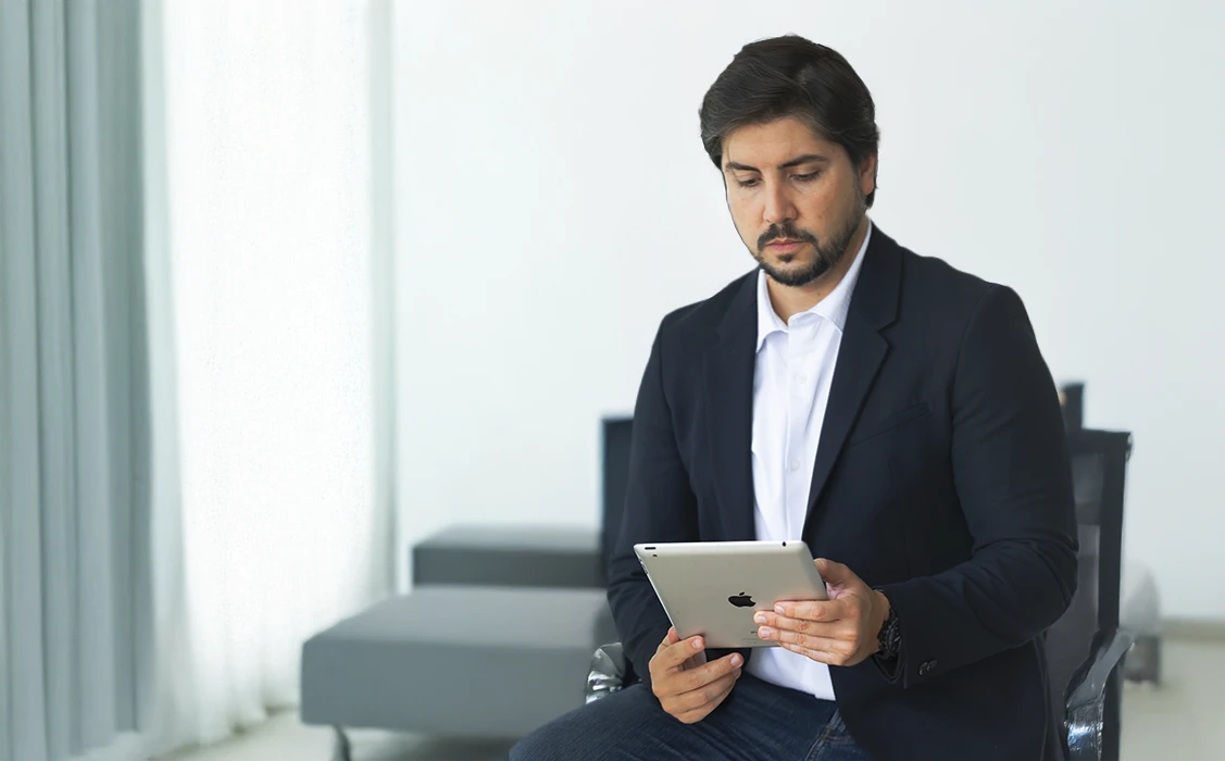 Homem de negócios com barba, cabelo escuro e roupa formal usando um tablet em ambiente corporativo moderno, transmitindo profissionalismo e tecnologia.